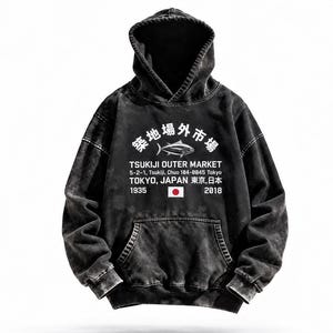 May include: A black acid-wash hooded sweatshirt featuring a white graphic on the chest. The print displays a fish icon, the text Tsukiji Outer Market, 5-2-1, Tsukiji, Chuo 104-0045 Tokyo, Tokyo, Japan, 1935, 2018, and a small Japanese flag.