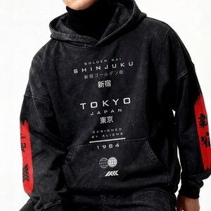 May include: Black hoodie with white text reading "GOLDEN GAI SHINJUKU 新宿 TOKYO JAPAN 東京 DESIGNED BY ALIENS 1984". Red accents with Japanese text adorn the sleeves. Features a front pocket and drawstring hood.