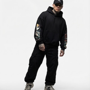 May include: Black hoodie with colorful sleeve art, paired with black cargo pants and a black baseball cap. The cap features a white logo. The outfit is completed with gray and beige sneakers.