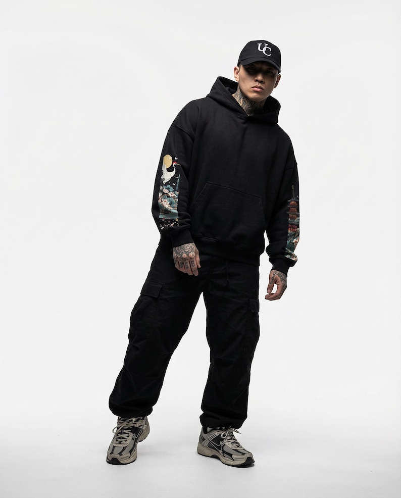 May include: Black hoodie with colorful sleeve art, paired with black cargo pants and a black baseball cap. The cap features a white logo. The outfit is completed with gray and beige sneakers.