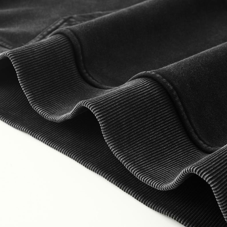 May include: A charcoal black sweatshirt featuring a thick, ribbed knit hem and cuffs. The fabric has a soft, slightly faded texture, showcasing the detailed stitching along the seams where the ribbed trim meets the main body of the garment.