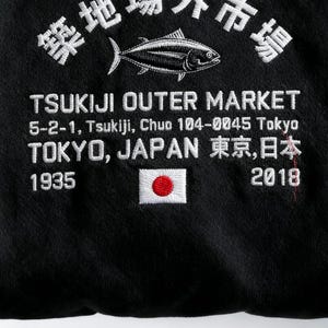 May include: A black hooded sweatshirt featuring white embroidered text that reads Tsukiji Outer Market, 5-2-1, Tsukiji, Chuo 104-0045 Tokyo, Tokyo, Japan, 1935, and 2018. The design includes a white fish graphic and a small embroidered Japanese flag.