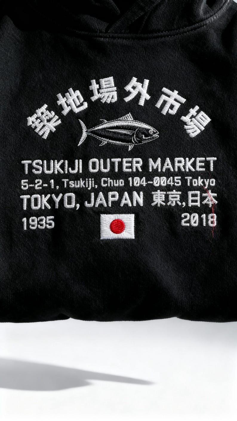 May include: A black hooded sweatshirt featuring white embroidered text that reads Tsukiji Outer Market, 5-2-1, Tsukiji, Chuo 104-0045 Tokyo, Tokyo, Japan, 1935, and 2018. The design includes a white fish graphic and a small embroidered Japanese flag.