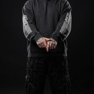 May include: Dark gray hoodie with the words "GRAND CULTURE" and "UNDR GRND" printed in white on the sleeves. The hoodie has a drawstring hood and a front pocket. The person is wearing black cargo pants.