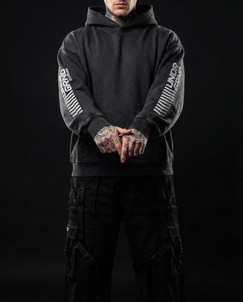 May include: Dark gray hoodie with the words "GRAND CULTURE" and "UNDR GRND" printed in white on the sleeves. The hoodie has a drawstring hood and a front pocket. The person is wearing black cargo pants.