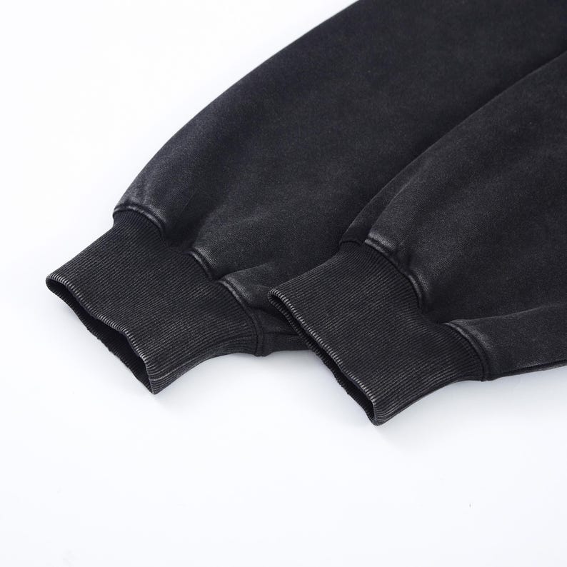 May include: Close-up of two black sweatshirt sleeves with ribbed cuffs. The fabric has a faded, vintage appearance. The cuffs are a darker shade of black than the sleeves.