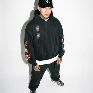 Pu&ograve; includere: Una persona indossa una felpa con cappuccio nera con dettagli floreali e testo giapponese, un berretto da baseball nero con logo bianco e pantaloni neri. La felpa ha una tasca frontale e una maglietta bianca. La persona indossa anche scarpe da ginnastica grigie.
