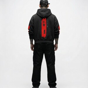 May include: A person wearing a black hoodie with red accents and Japanese text on the back, paired with black cargo trousers and a black cap. The hoodie has red paint strokes on the sleeves.