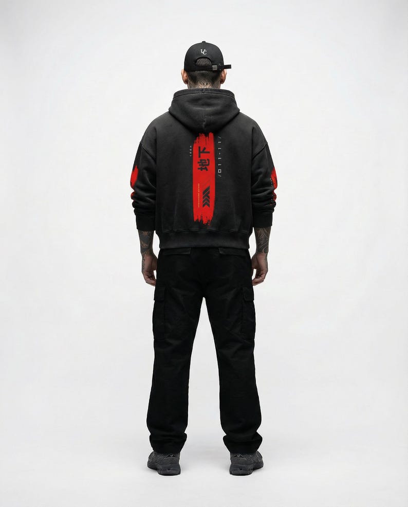 May include: A person wearing a black hoodie with red accents and Japanese text on the back, paired with black cargo trousers and a black cap. The hoodie has red paint strokes on the sleeves.