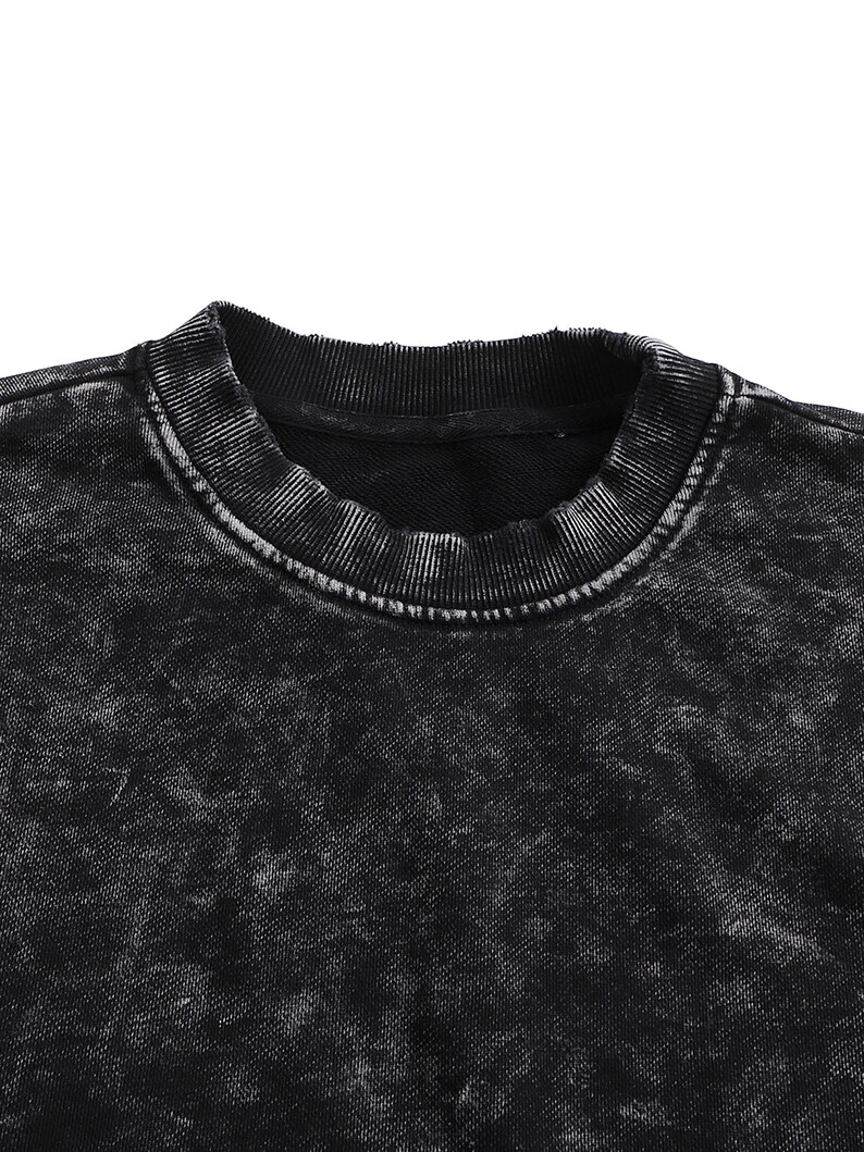 May include: Close-up of a black crew neck sweatshirt with a distressed, acid wash effect. The collar and neckline are ribbed, and the fabric has a textured appearance. The garment is a dark, neutral color.