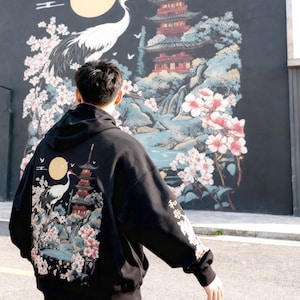 May include: Black hoodie with a Japanese-inspired design featuring a crane, pagoda, and cherry blossoms. The back of the hoodie displays a detailed illustration with a full moon and Japanese text on the sleeve.