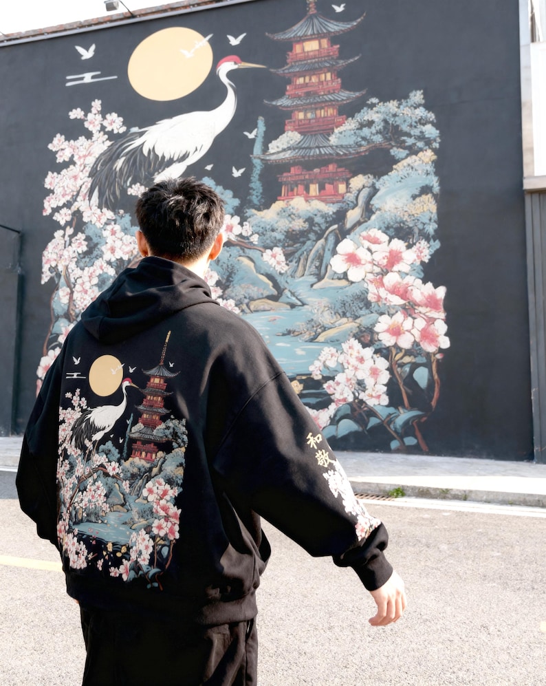 May include: Black hoodie with a Japanese-inspired design featuring a crane, pagoda, and cherry blossoms. The back of the hoodie displays a detailed illustration with a full moon and Japanese text on the sleeve.