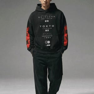 May include: Black hoodie and trousers set. The hoodie displays the text "SHINJUKU TOKYO JAPAN" in white, with red detailing on the sleeves. The year "1984" is also printed on the hoodie. The model is wearing black trainers.