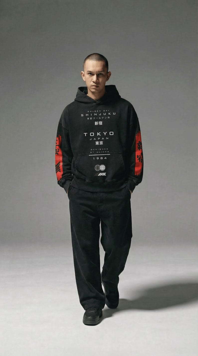 May include: Black hoodie and trousers set. The hoodie displays the text "SHINJUKU TOKYO JAPAN" in white, with red detailing on the sleeves. The year "1984" is also printed on the hoodie. The model is wearing black trainers.