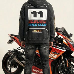 May include: A person wearing a black hoodie with the text "11 ELEVEN RACE CLUB" and a motorcycle helmet stands in front of a red and black Suzuki motorcycle. The motorcycle has the text "GX 1000" and "UCR". The person is wearing black jeans.