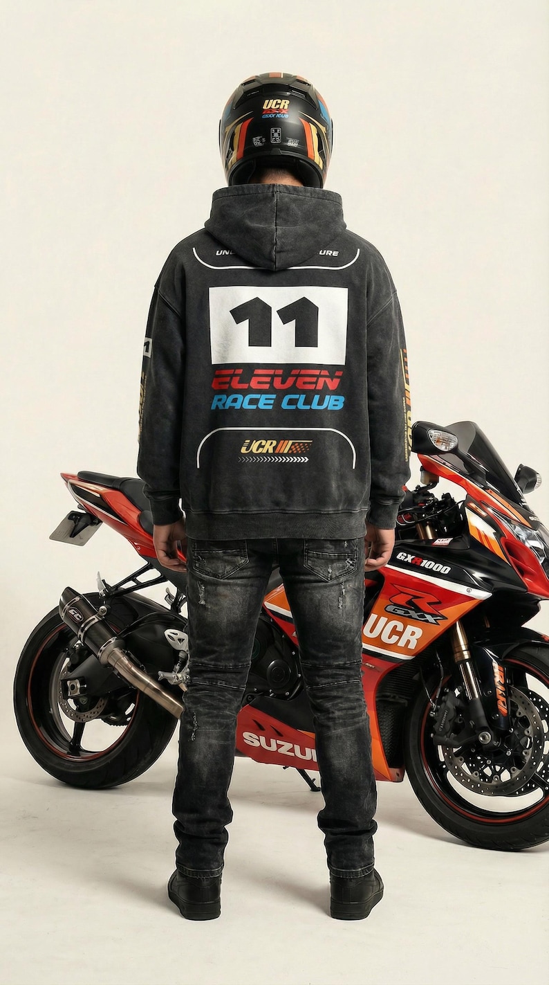 May include: A person wearing a black hoodie with the text "11 ELEVEN RACE CLUB" and a motorcycle helmet stands in front of a red and black Suzuki motorcycle. The motorcycle has the text "GX 1000" and "UCR". The person is wearing black jeans.