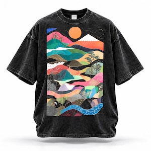 May include: A black acid-wash t-shirt featuring a large rectangular graphic print of a stylized landscape. The design includes rolling hills in shades of pink, green, and blue, a white-capped mountain, a bright orange sun, and a blue river.