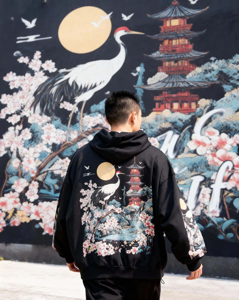 May include: Black hoodie featuring a Japanese-inspired design with a crane, pagoda, and cherry blossoms. The artwork includes a full moon and is set against a dark background. The sleeves also have the same design.