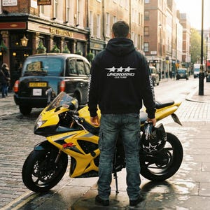 May include: A yellow and black Suzuki GSX-R motorcycle parked on a wet cobblestone street. A person wearing a black hoodie with "UNDRGRND CULTURE" text stands near the motorcycle, holding a helmet. A black taxi is in the background.