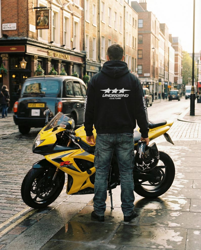 May include: A yellow and black Suzuki GSX-R motorcycle parked on a wet cobblestone street. A person wearing a black hoodie with "UNDRGRND CULTURE" text stands near the motorcycle, holding a helmet. A black taxi is in the background.