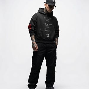 May include: A person wearing a black hoodie with "TOKYO JAPAN" text and a black baseball cap. The hoodie has red accents on the sleeves and a front pocket. The person is also wearing black trousers and shoes.