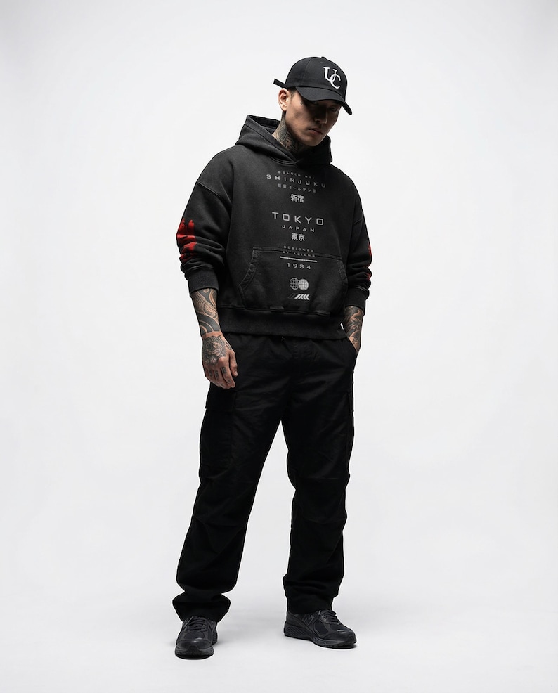 May include: A person wearing a black hoodie with "TOKYO JAPAN" text and a black baseball cap. The hoodie has red accents on the sleeves and a front pocket. The person is also wearing black trousers and shoes.
