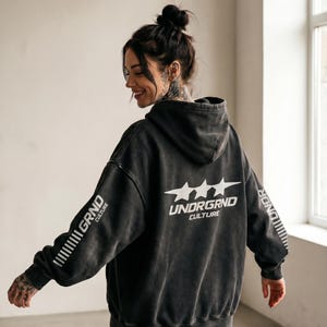 May include: A dark gray hoodie with the text "UNDRGRND CULTURE" and three stars on the back. The left sleeve has the text "GRND CULTURE" printed vertically. The hoodie is worn by a person with tattoos and a bun hairstyle.