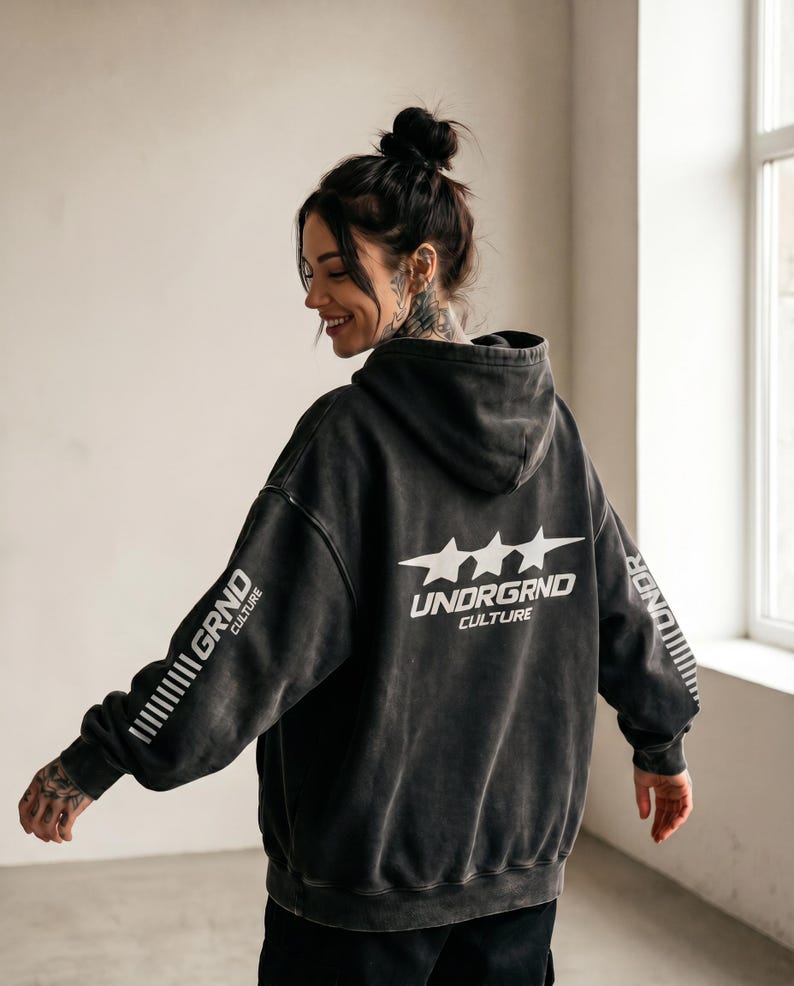 May include: A dark gray hoodie with the text "UNDRGRND CULTURE" and three stars on the back. The left sleeve has the text "GRND CULTURE" printed vertically. The hoodie is worn by a person with tattoos and a bun hairstyle.