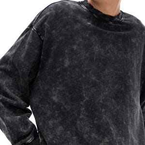May include: A black, acid-wash style sweatshirt with a crew neck and long sleeves. The fabric has a mottled, faded appearance, with lighter gray patches against a darker black base. The cuffs and neckline are ribbed.