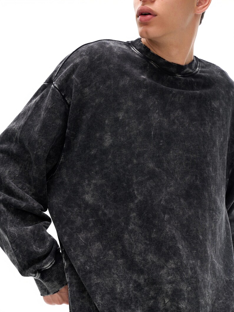 May include: A black, acid-wash style sweatshirt with a crew neck and long sleeves. The fabric has a mottled, faded appearance, with lighter gray patches against a darker black base. The cuffs and neckline are ribbed.