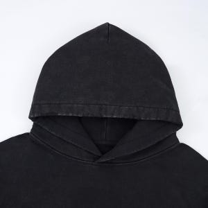 May include: A close-up of a black hoodie with a drawstring neckline and a large hood. The fabric has a slightly faded appearance, suggesting a vintage or washed style. The hoodie is made of a soft material.