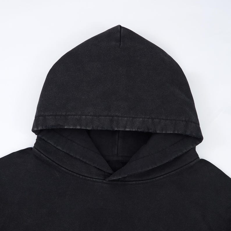 May include: A close-up of a black hoodie with a drawstring neckline and a large hood. The fabric has a slightly faded appearance, suggesting a vintage or washed style. The hoodie is made of a soft material.