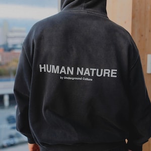May include: A black hooded sweatshirt featuring the text HUMAN NATURE by Underground Culture printed in white on the back. The garment has a hood, long sleeves, and a ribbed hem, showcasing a faded, washed-out black fabric finish throughout.