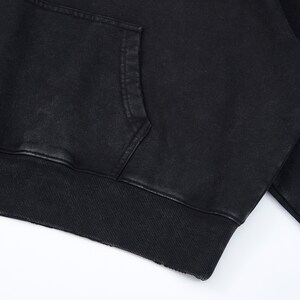 May include: Close-up of a black hoodie with a kangaroo pocket. The hoodie features a ribbed hem and cuffs. The fabric has a slightly faded, textured appearance. The garment is made of a soft material.