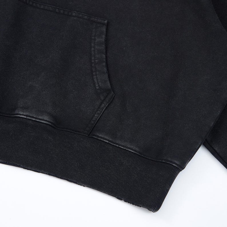 May include: Close-up of a black hoodie with a kangaroo pocket. The hoodie features a ribbed hem and cuffs. The fabric has a slightly faded, textured appearance. The garment is made of a soft material.