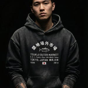 May include: A charcoal grey hooded sweatshirt featuring a white graphic print of a fish, the text Tsukiji Outer Market, 5-2-1, Tsukiji, Chuo 104-0845 Tokyo, Tokyo, Japan, 1935, 2018, and a small Japanese flag icon centered on the front chest.