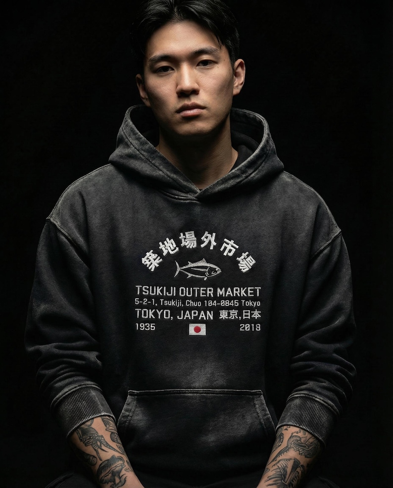 May include: A charcoal grey hooded sweatshirt featuring a white graphic print of a fish, the text Tsukiji Outer Market, 5-2-1, Tsukiji, Chuo 104-0845 Tokyo, Tokyo, Japan, 1935, 2018, and a small Japanese flag icon centered on the front chest.