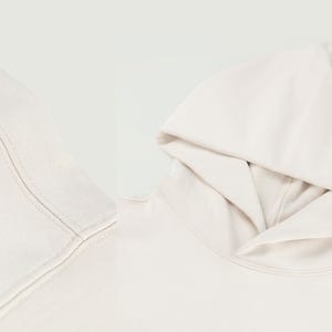 Pu&ograve; includere: Primo piano di una felpa con cappuccio color crema, che mostra il cappuccio e lo scollo. Il tessuto appare morbido e le cuciture sono visibili. La felpa con cappuccio &egrave; di un colore neutro, adatta a vari outfit.