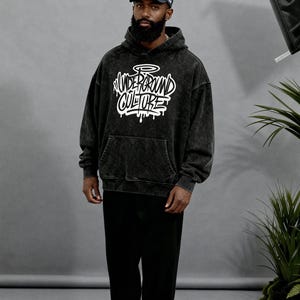 May include: A black acid-wash hooded sweatshirt featuring the white text "UNDERGROUND CULTURE" in a graffiti-style font on the chest, paired with black trousers, a white and black baseball cap, and grey and black athletic sneakers.