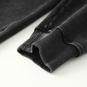 May include: A long-sleeved garment sleeve featuring a dark charcoal gray, acid-washed fabric finish. The sleeve ends in a thick, ribbed knit cuff that matches the dark gray color of the main fabric, providing a textured and fitted appearance.
