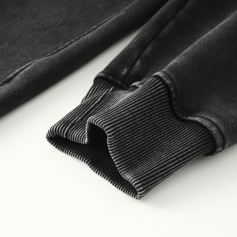 May include: A long-sleeved garment sleeve featuring a dark charcoal gray, acid-washed fabric finish. The sleeve ends in a thick, ribbed knit cuff that matches the dark gray color of the main fabric, providing a textured and fitted appearance.