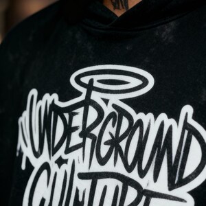 May include: A black hooded sweatshirt with a washed, faded texture. The front features large, white, graffiti-style lettering that reads "UNDERGROUND CULTURE" with a halo graphic above the text. The design has a dripping paint effect.