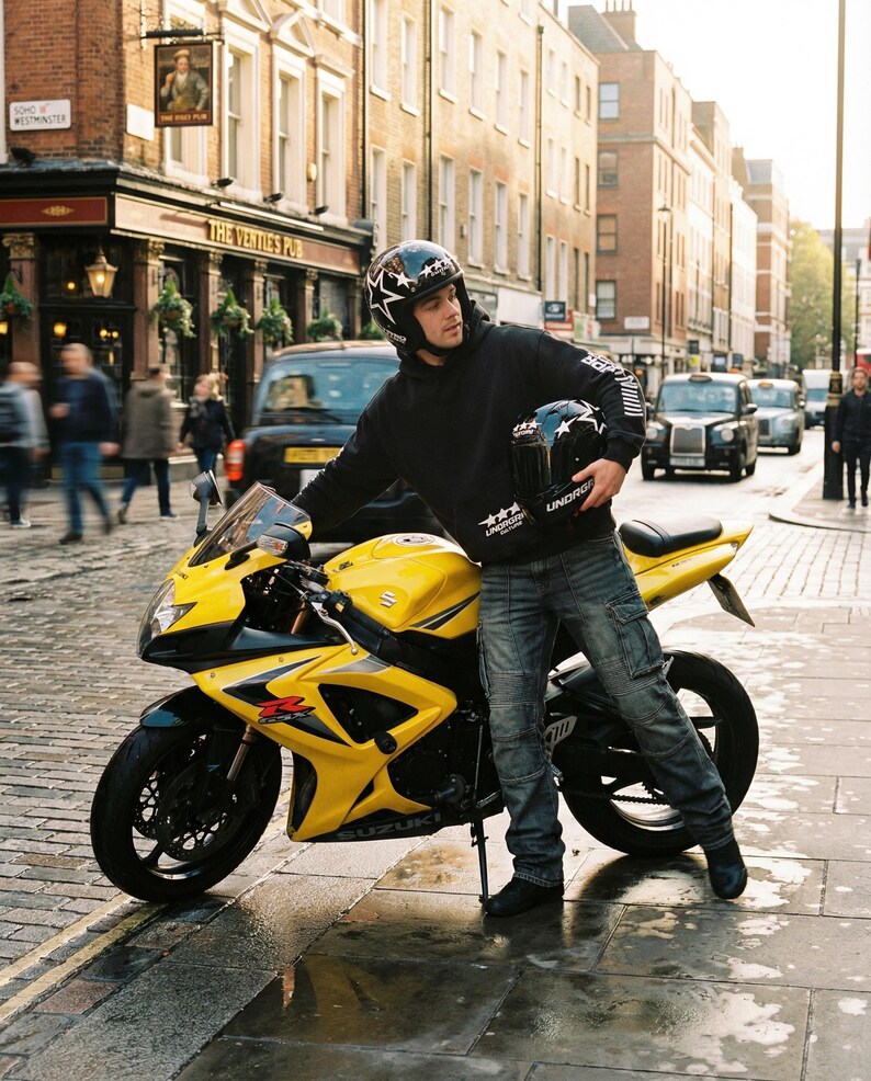 May include: A yellow Suzuki GSX-R motorcycle parked on a wet cobblestone street. The motorcycle has black accents and a black tire. A person wearing a black helmet with star designs and a black hoodie stands next to the motorcycle.