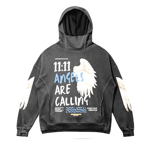 May include: Dark gray hoodie with a hood and long sleeves. The front features the text "11:11 Angels Are Calling" with angel wing graphics. The sleeves also have angel wing graphics. The hoodie has the text "UNDERGROUND CULTURE" and "RAISE YOUR VIBRATION".