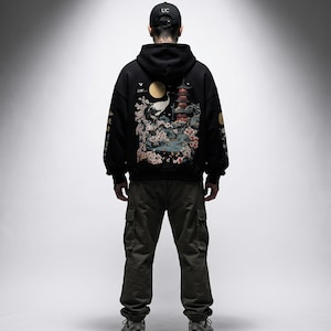 May include: A person wearing a black hoodie with a Japanese-inspired design on the back, featuring a crane, pagoda, and cherry blossoms. The person is also wearing olive green cargo pants and a black baseball cap with "UC" on it.