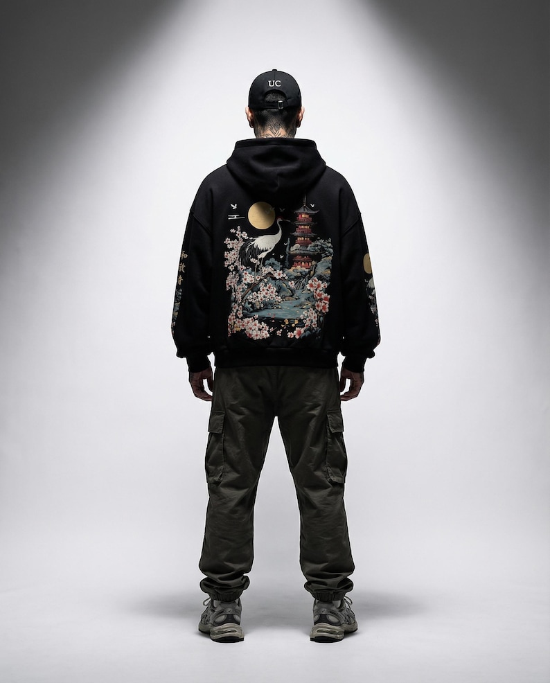 May include: A person wearing a black hoodie with a Japanese-inspired design on the back, featuring a crane, pagoda, and cherry blossoms. The person is also wearing olive green cargo pants and a black baseball cap with "UC" on it.