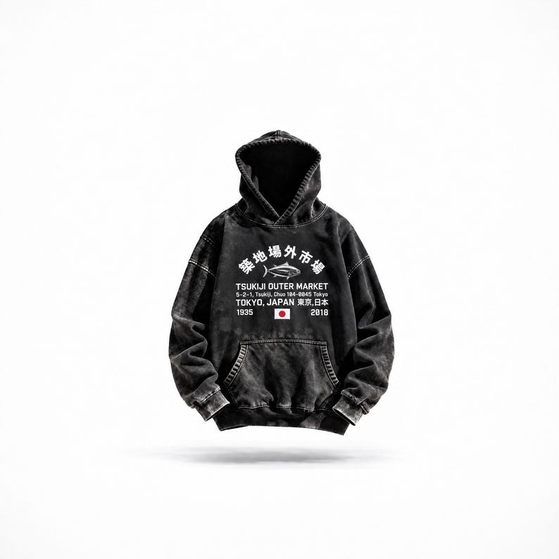 May include: A black acid-wash hooded sweatshirt featuring a white graphic on the chest. The print displays a fish icon, the text Tsukiji Outer Market, 5-2-1, Tsukiji, Chuo 104-0045 Tokyo, Tokyo, Japan, 1935, 2018, and a small Japanese flag.