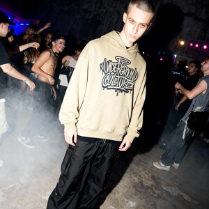 May include: A beige hooded sweatshirt featuring a black graphic print on the chest with the text "UNDERGROUND CULTURE" in a stylized graffiti font. The hoodie includes a front kangaroo pocket, long sleeves with ribbed cuffs, and a drawstring hood.