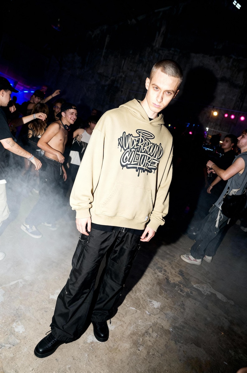 May include: A beige hooded sweatshirt featuring a black graphic print on the chest with the text "UNDERGROUND CULTURE" in a stylized graffiti font. The hoodie includes a front kangaroo pocket, long sleeves with ribbed cuffs, and a drawstring hood.