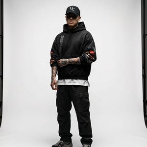 Pu&ograve; includere: Una persona indossa una felpa con cappuccio nera con un design grafico sulle maniche, pantaloni cargo neri, un berretto da baseball nero con scritte bianche e scarpe da ginnastica grigie. La felpa ha del testo sulla manica sinistra.